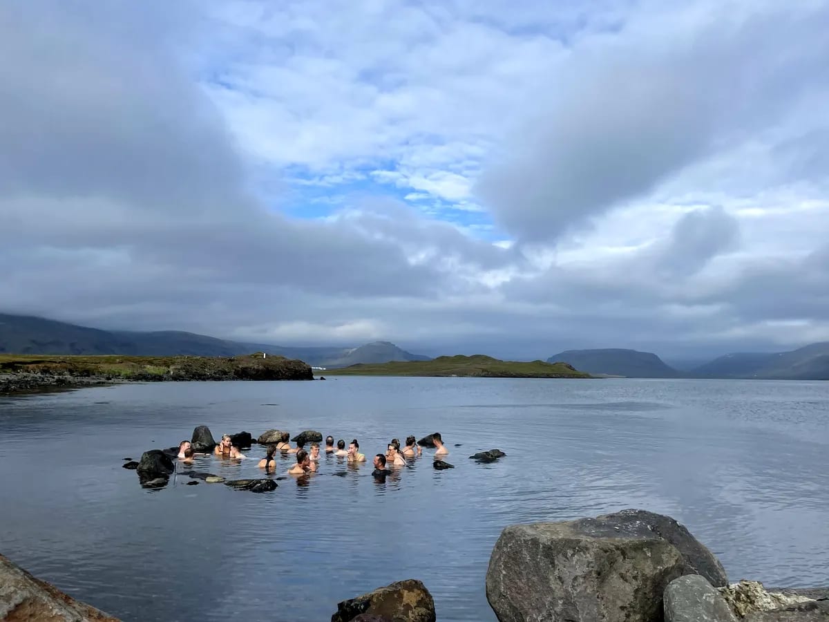 Guests soaking in a natural geothermal sea pool at Hvammsvík, surrounded by black volcanic rocks, calm fjord water and Icelandic mountains