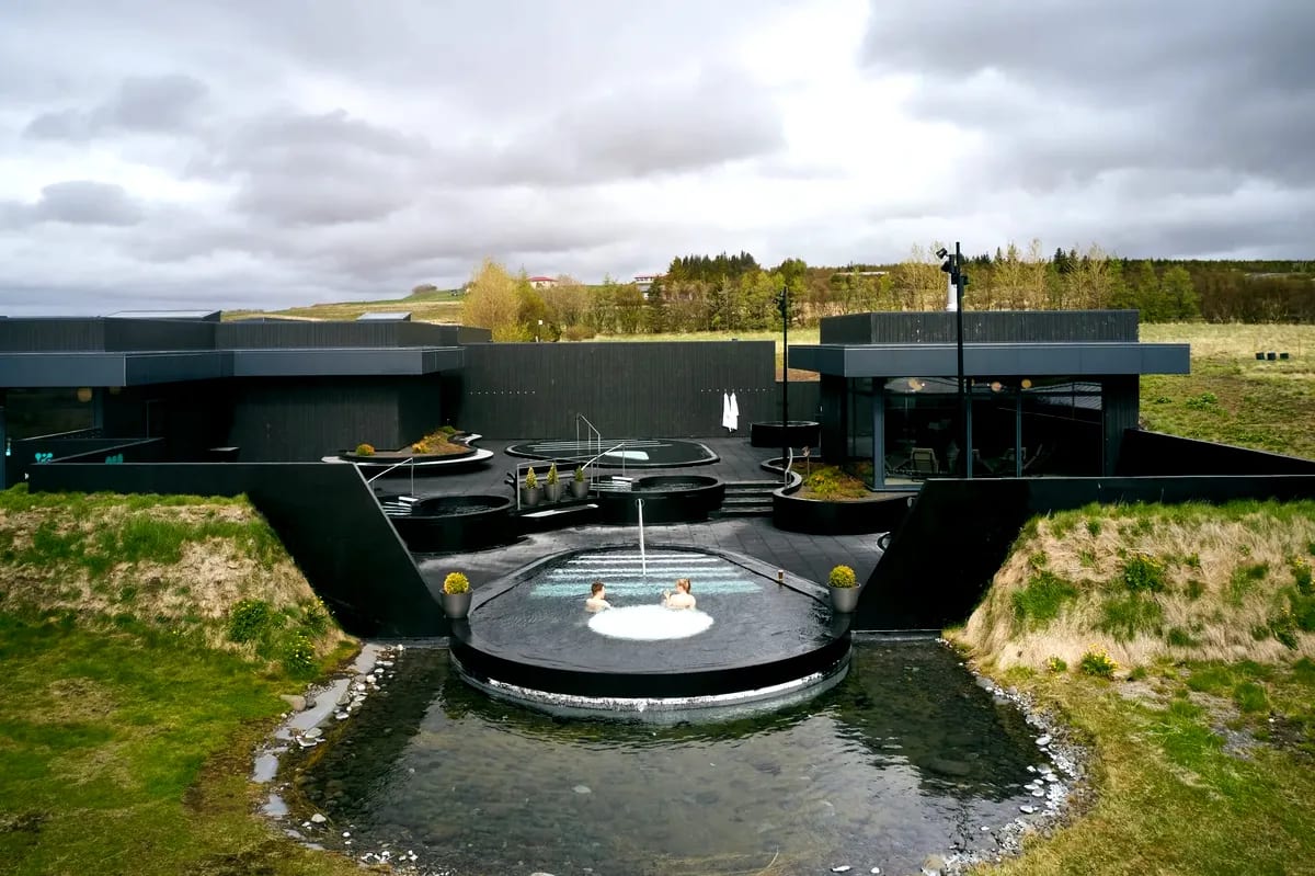 Aerial view of Krauma Spa in Iceland with outdoor geothermal hot tubs, modern black architecture, green hills and two guests relaxing