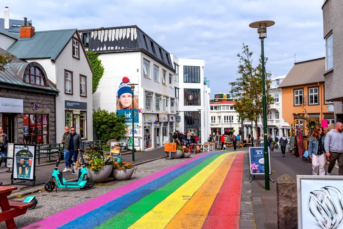 Rainbow-painted street in downtown Reykjavik with shops, scooters, flower planters and travelers under cloudy May skies in Iceland