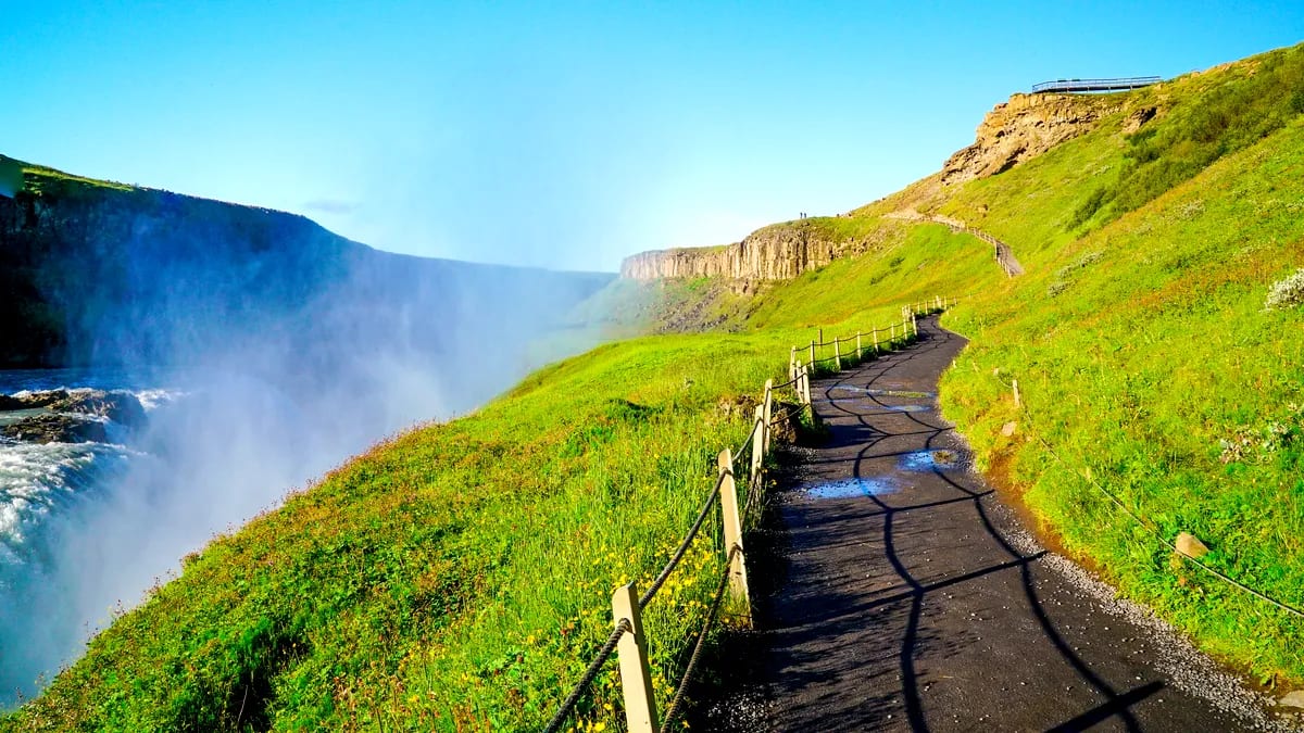 Misty Iceland waterfall beside a winding cliffside trail, lush green hills and bright May sunshine on a scenic nature walk in Iceland