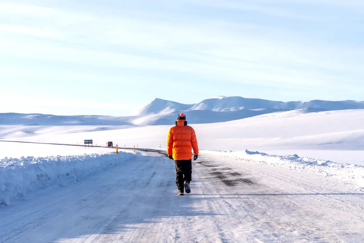 Traveler in orange winter jacket walking along a snowy Iceland road toward white mountains under pale May light and clear blue sky