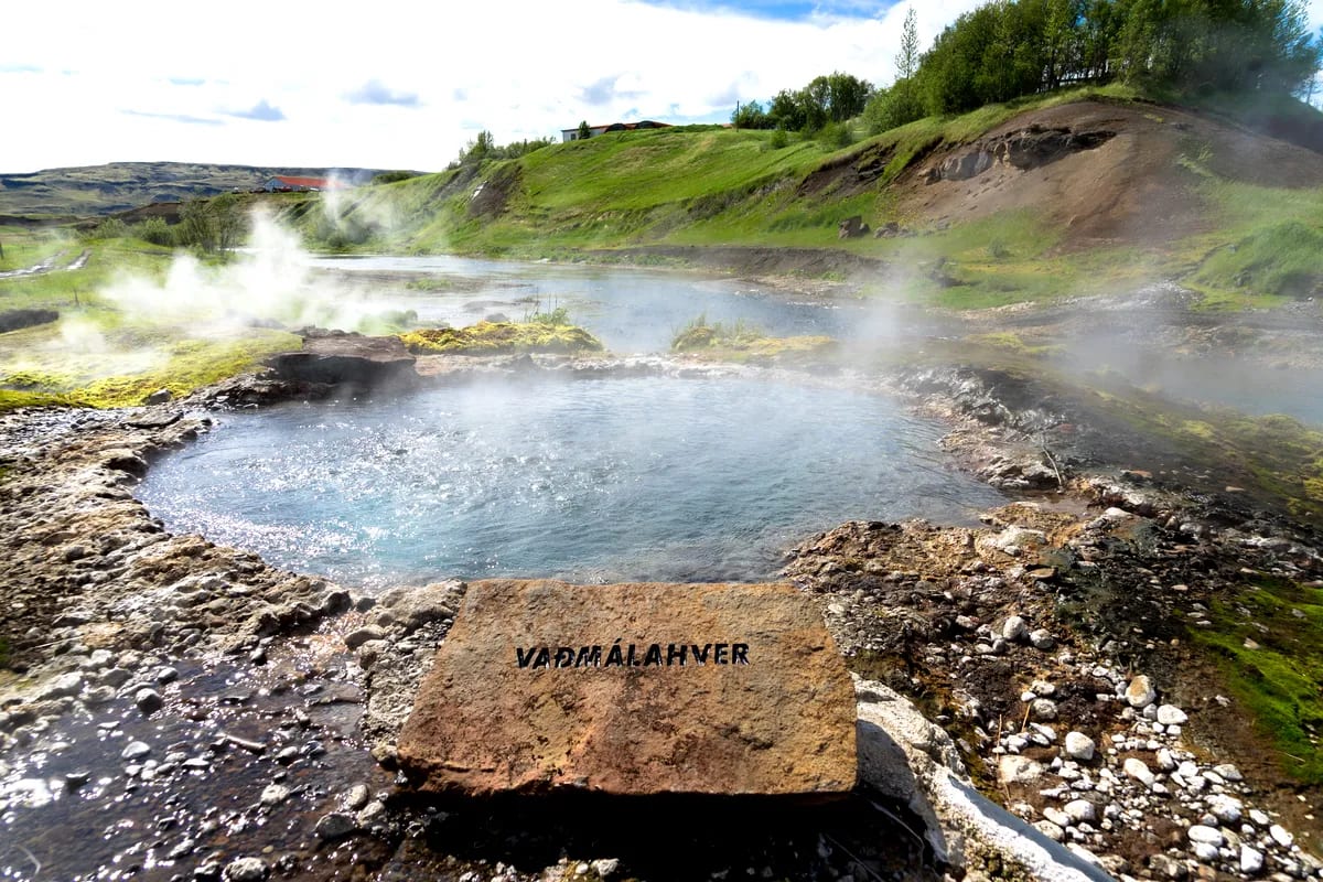 Steaming Icelandic geothermal hot spring at Vaðmálahver with blue water, rocky edges and bright green hills on a sunny May day
