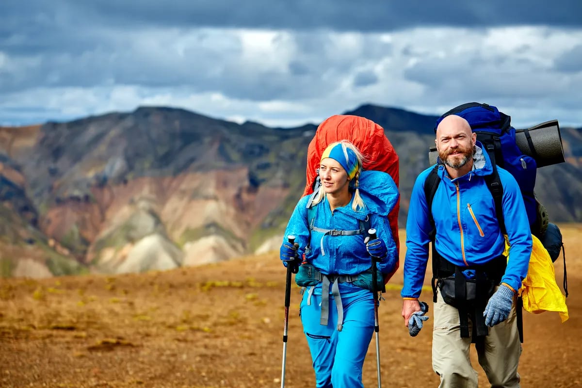 Two backpackers hiking in Iceland’s colorful June highlands with trekking poles, blue jackets, heavy packs, and cloudy mountains behind.