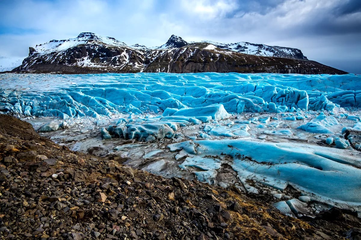 Bright blue glacier in Iceland in June with cracked ice, rocky moraine, snow-capped mountains, and dramatic clouds in summer light.
