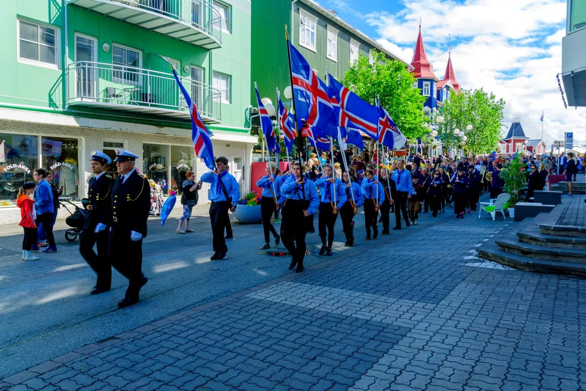 Icelandic National Day parade in June with flag bearers marching through a colorful town street under sunny skies and red church spires.