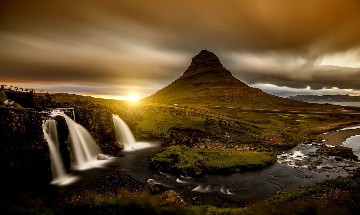 Kirkjufellsfoss waterfall cascading below Kirkjufell mountain at golden sunset, with storm clouds, mossy lava rocks and river views