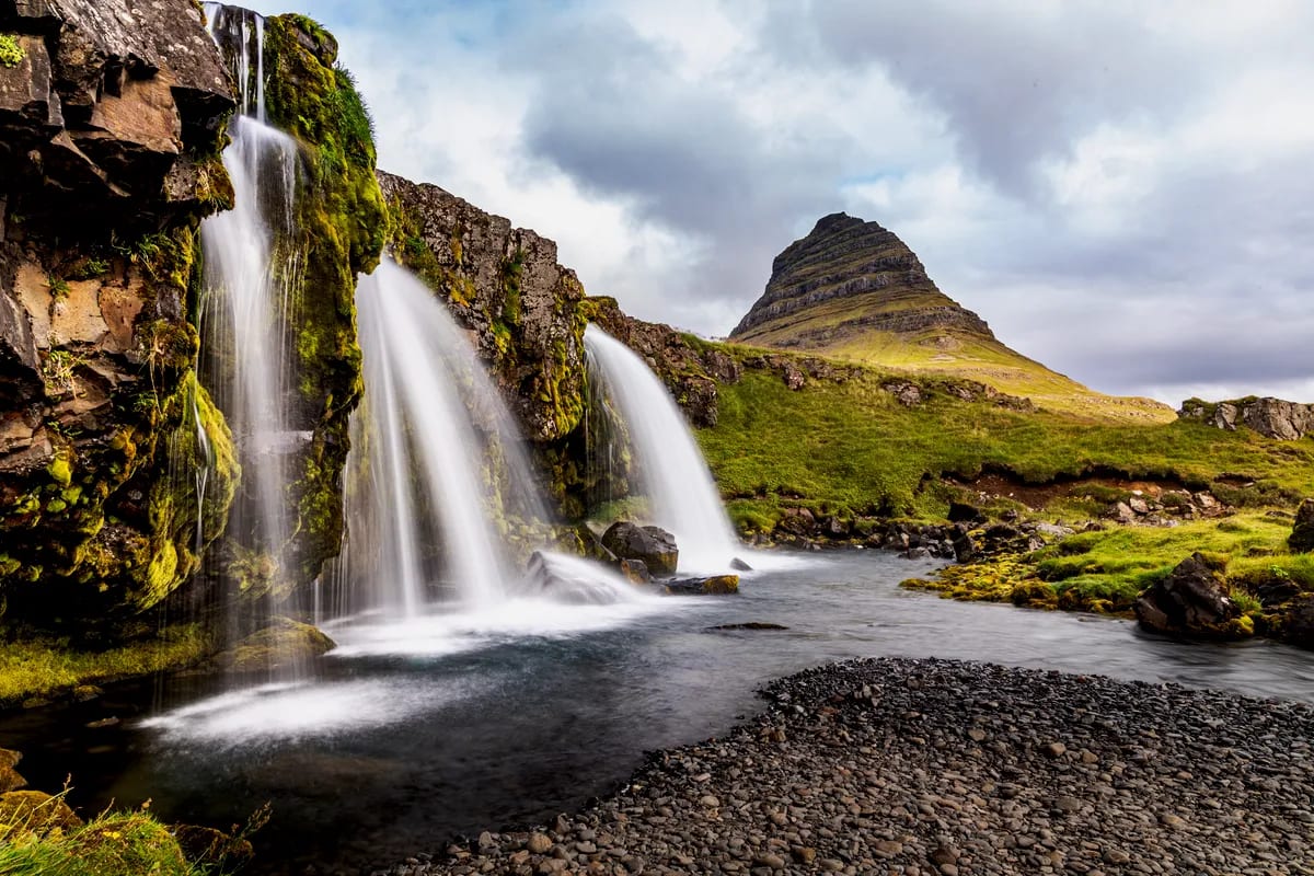 Close-up of Kirkjufellsfoss waterfall beside moss-covered lava cliffs, with Kirkjufell mountain rising under cloudy Iceland skies