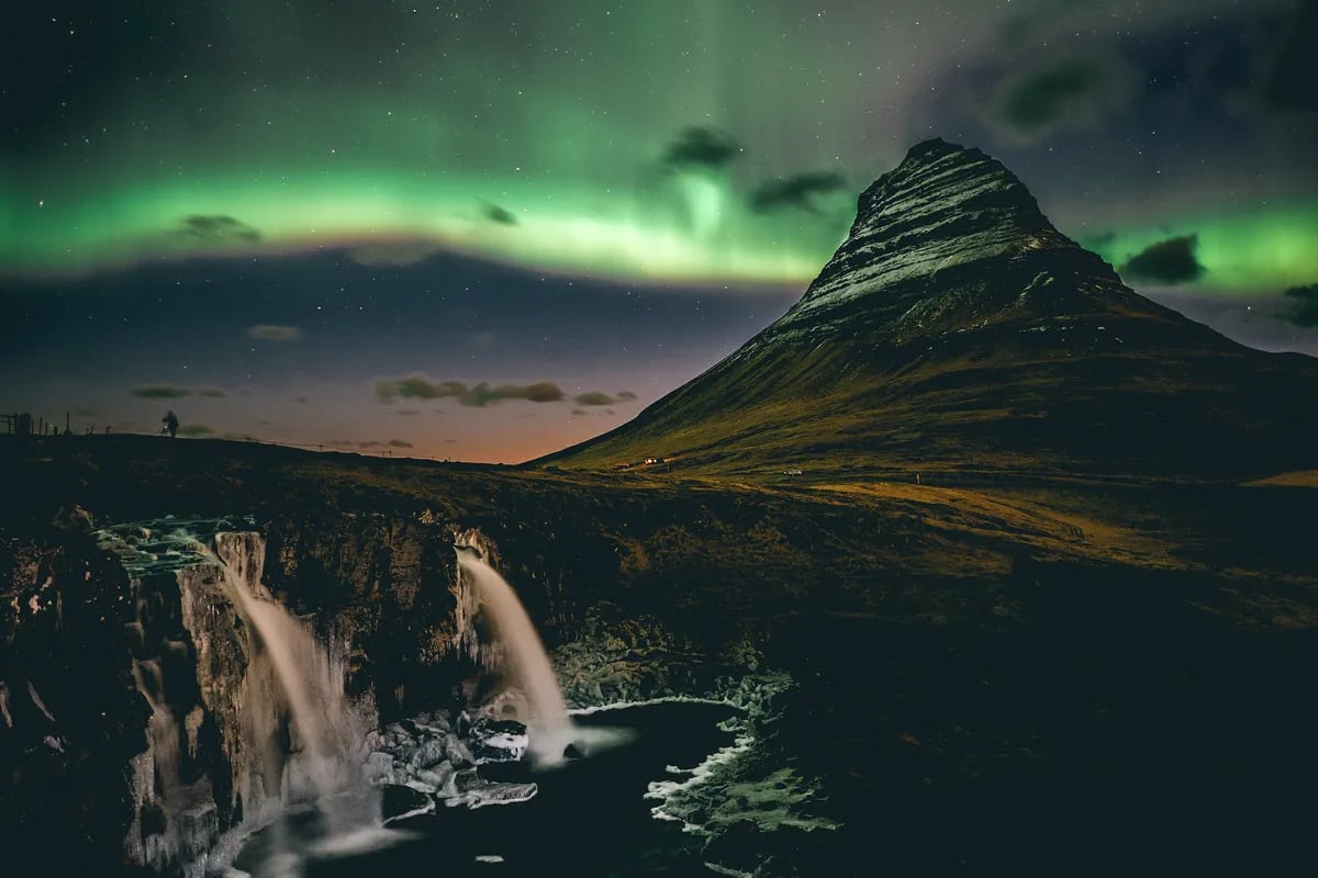 Northern lights glow over Kirkjufell mountain and Kirkjufellsfoss waterfall in Iceland, with stars, ice, and dramatic April night scenery