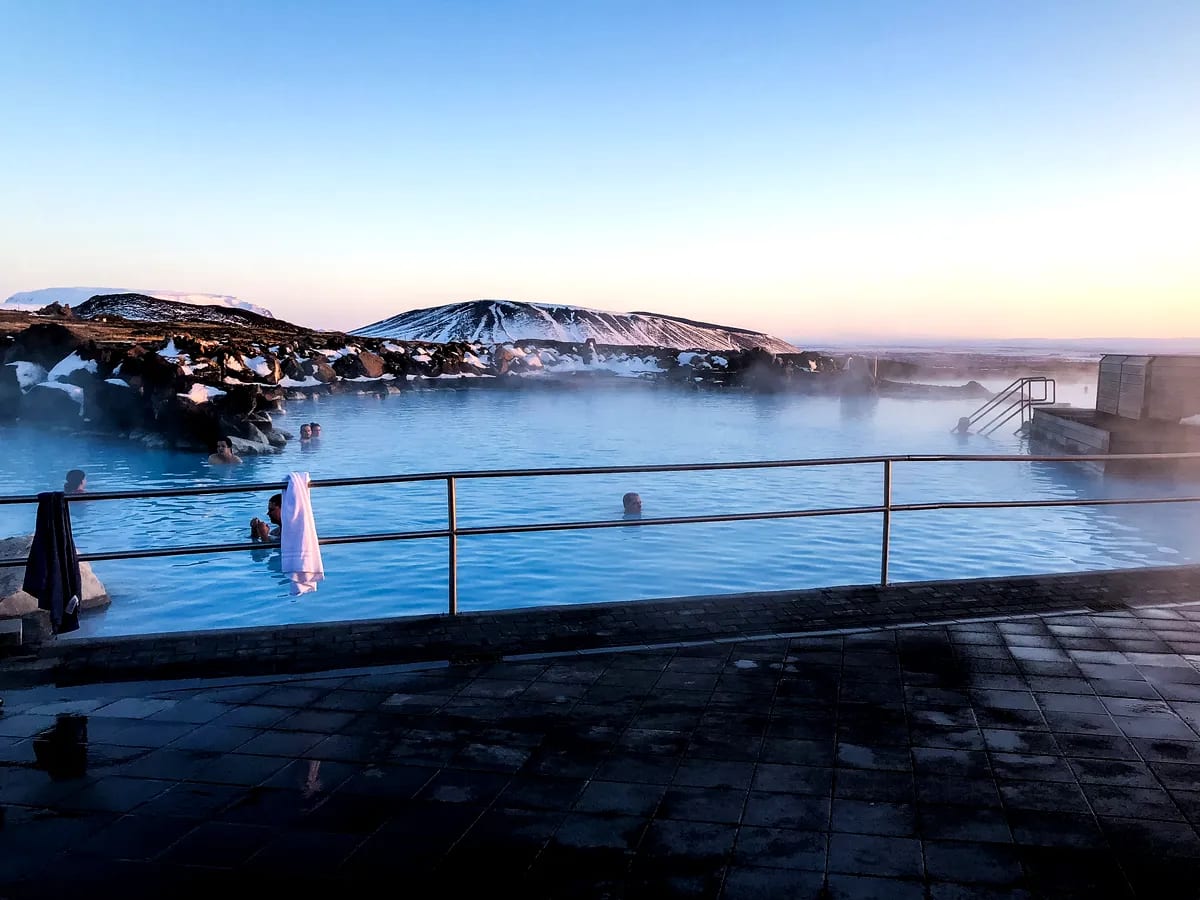 People relax in a steaming blue geothermal lagoon in Iceland, with snow-dusted hills, lava rocks, and soft April sunrise light