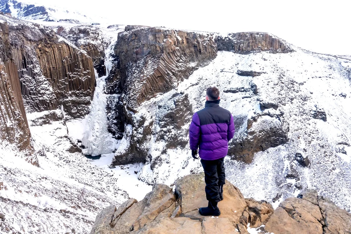 Traveler in purple jacket overlooking snowy Litlanesfoss waterfall and basalt columns on the winter hike to Hengifoss in East Iceland