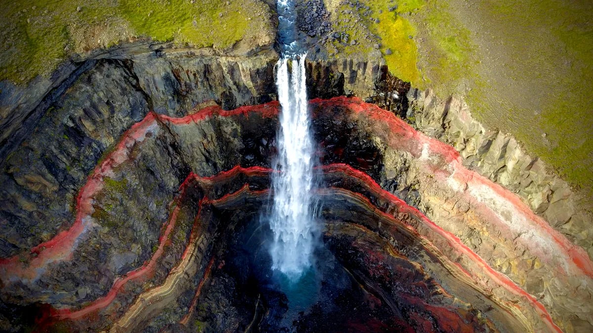 Aerial view of Hengifoss Waterfall plunging into a canyon with vivid red clay layers, basalt cliffs and green Icelandic highlands