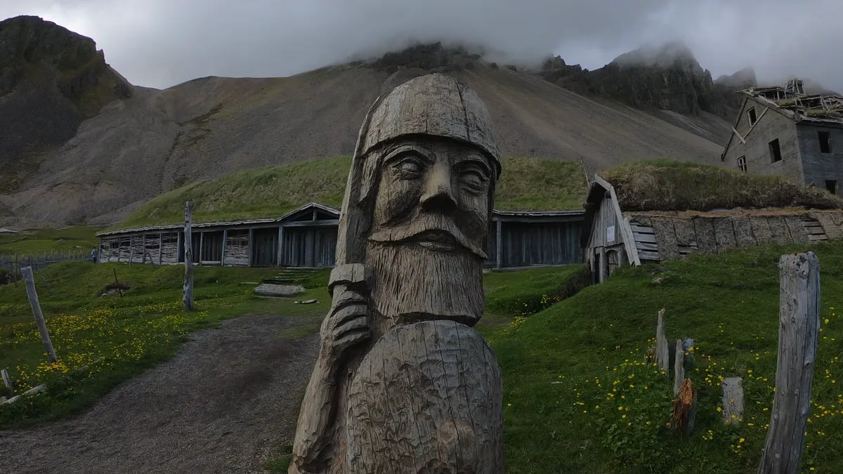 Carved wooden Viking statue stands before Icelandic turf-roof buildings and misty mountains, inviting visitors into a temporary Viking experience