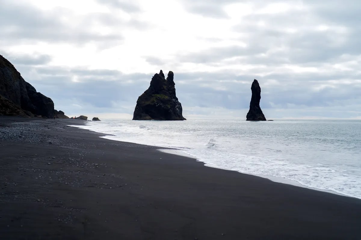 Black sand beach in Iceland with dark volcanic shoreline, gentle Atlantic waves and dramatic Reynisdrangar sea stacks under cloudy sky