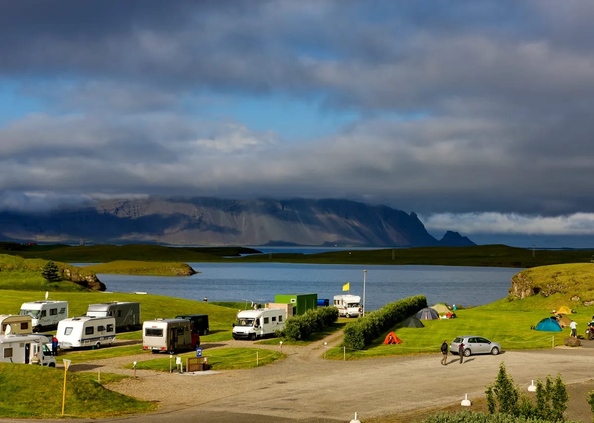 Camper vans and tents at a lakeside Iceland campground under dramatic clouds, with mountains across the water in February travel scene