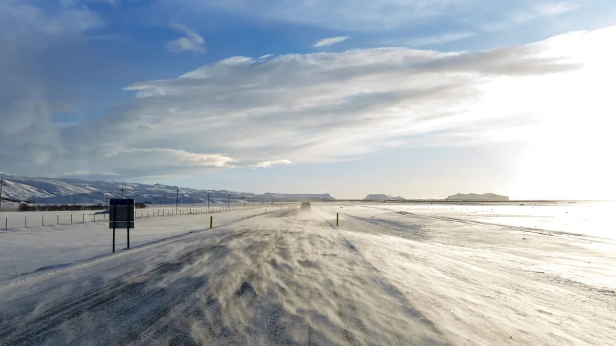 Snow blowing across an icy Iceland road in February, with bright winter sun, snowy fields and distant coastal mountains ahead