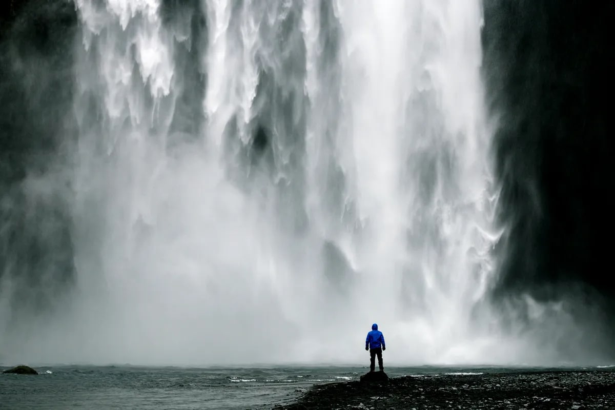Traveler in blue jacket stands before misty Skógafoss waterfall in Iceland, dwarfed by thunderous cascades and dark volcanic cliffs
