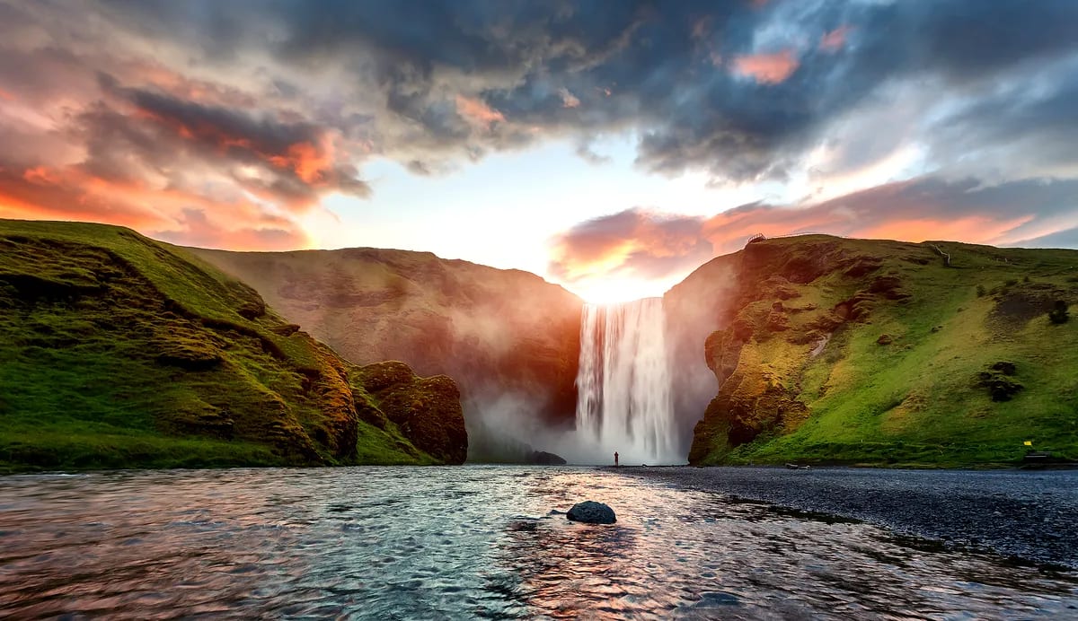 Skógafoss waterfall glowing at sunset between green Icelandic cliffs, with mist rising over river and lone traveler beneath dramatic sky