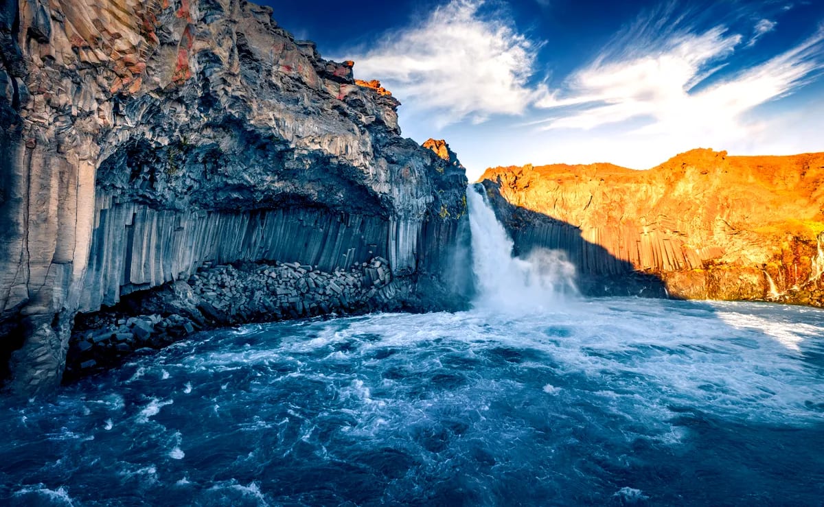 Aldeyjarfoss waterfall crashing into a blue glacial river between dramatic basalt columns and sunlit volcanic cliffs in North Iceland