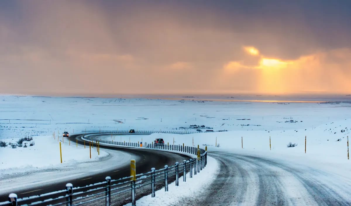 Winding icy road through snow-covered Iceland plains in December, with cars driving toward a golden winter sunset under stormy skies