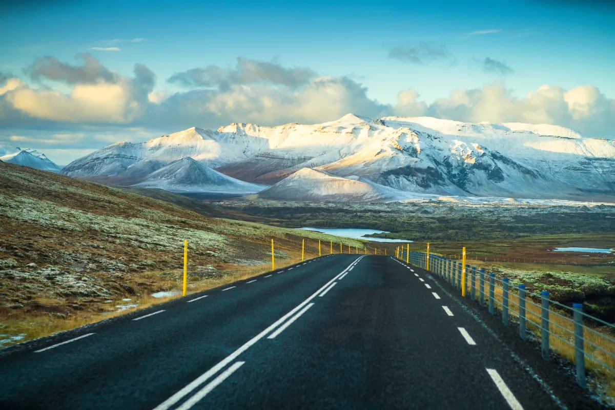 Iceland Ring Road stretching toward snow-covered mountains, yellow roadside posts and winter hills beneath a glowing blue sky