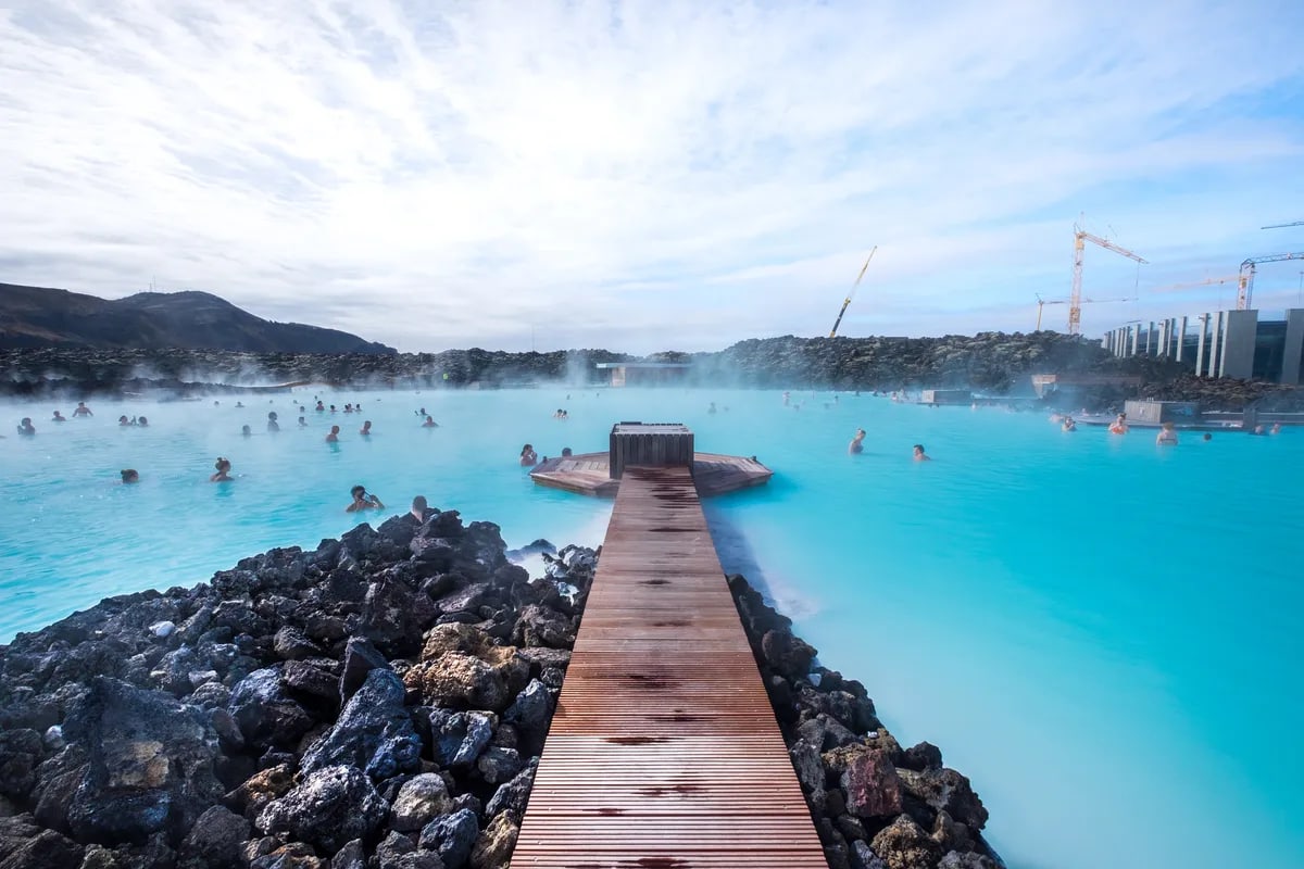 Blue Lagoon in Iceland with milky blue geothermal water, wooden walkway, black lava rocks and visitors relaxing in steaming pools