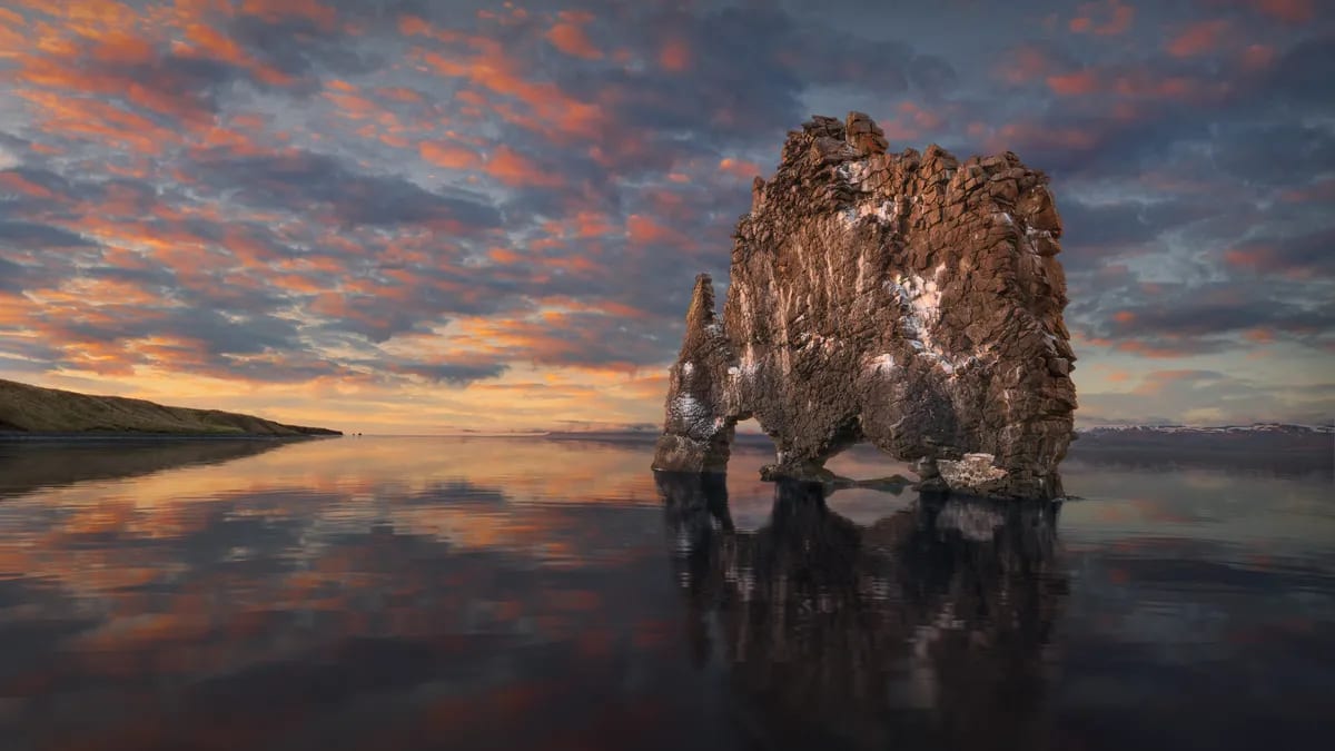 Hvítserkur sea stack on North Iceland’s coast at sunset, reflected in wet black sand with dramatic orange clouds over the bay