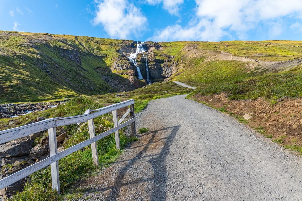 Gravel path to a hillside waterfall in East Iceland, with wooden fence, green mountain slopes and bright blue sky over the trail