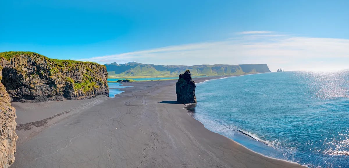 Reynisfjara black sand beach on Iceland’s South Coast with basalt cliffs, sea stack, blue ocean and distant Reynisdrangar rocks