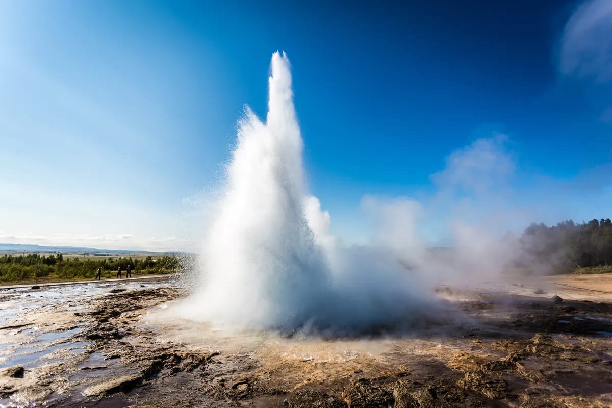 Strokkur geyser erupting on Iceland’s Golden Circle, with steaming geothermal field, tourists nearby and clear blue sky above