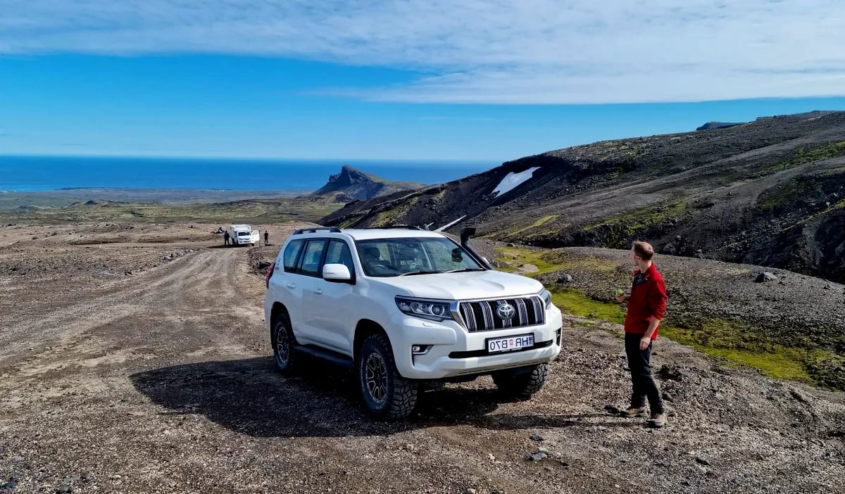 White 4x4 SUV on a rough gravel F-road in Iceland’s Highlands, with a traveler assessing terrain, volcanic slopes and ocean views