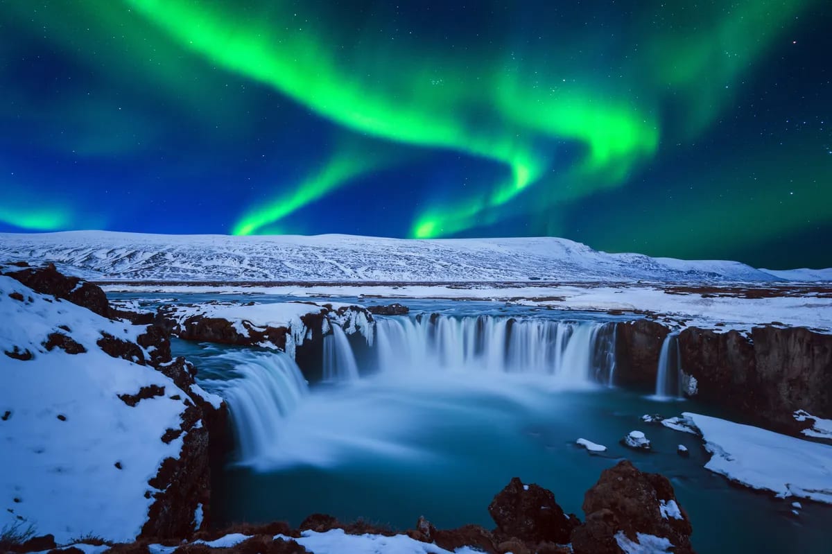 Goðafoss Waterfall in North Iceland under green northern lights, snowy cliffs, frozen river, and starry sky for location guide