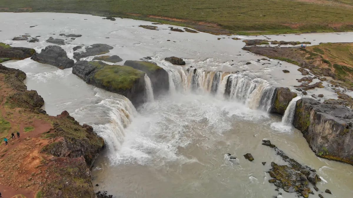 Aerial view of Goðafoss Waterfall in North Iceland, with powerful cascades, rocky cliffs, muddy river, hiking trails, and visitors