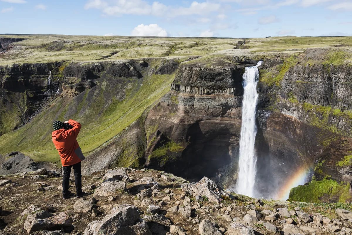 Traveler photographing Haifoss waterfall from a rocky cliff, with a rainbow in the mist and green Icelandic canyon walls below