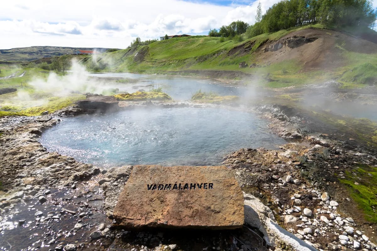Vaðmálahver hot spring at Iceland’s Secret Lagoon, with steaming mineral water, mossy rocks, green hills, and a bright geothermal landscape