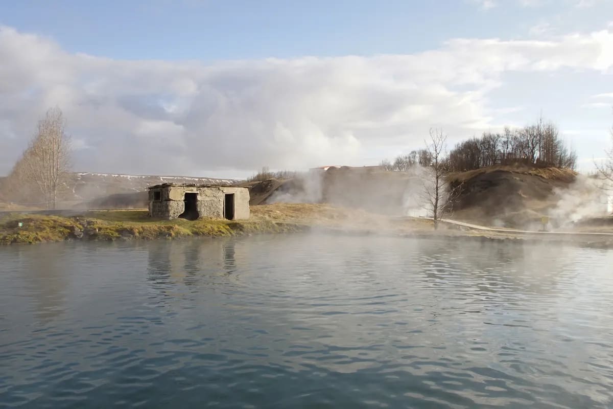 Steam rises over Iceland’s Secret Lagoon, reflecting an old stone bathhouse, bare trees, and hills around the warm geothermal pool