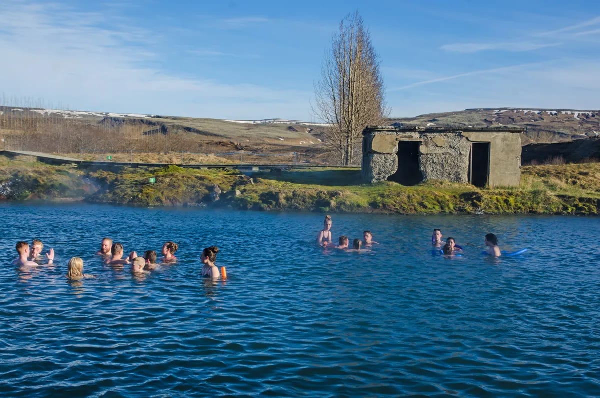 Visitors relaxing in Iceland’s Secret Lagoon, with steaming blue geothermal water, old stone hut, bare trees, and snowy hills under clear sky