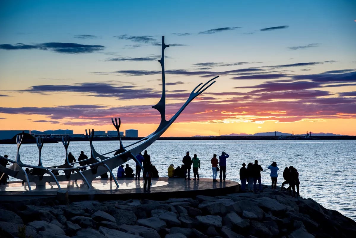 Visitors watch sunset beside the Sun Voyager sculpture on Reykjavik’s waterfront, with Faxaflói Bay and mountains glowing in the distance