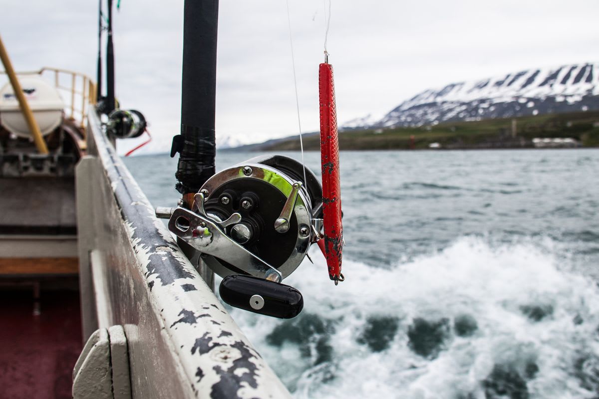 Close-up of fishing reel and red lure on a boat in Icelandic fjord waters, with snowy mountains and sea spray in the background