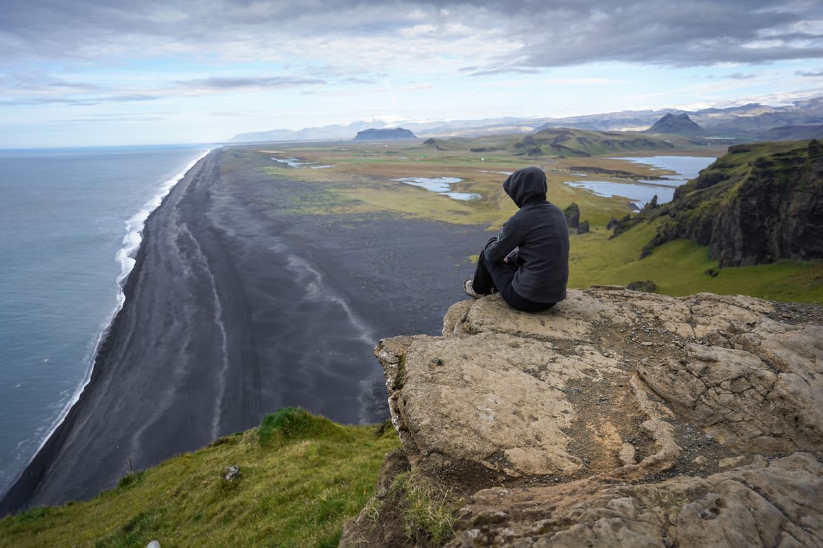 Traveler sits on a cliff above Reynisfjara black sand beach, overlooking Iceland’s dramatic coastline, ocean waves, cliffs, and lagoon
