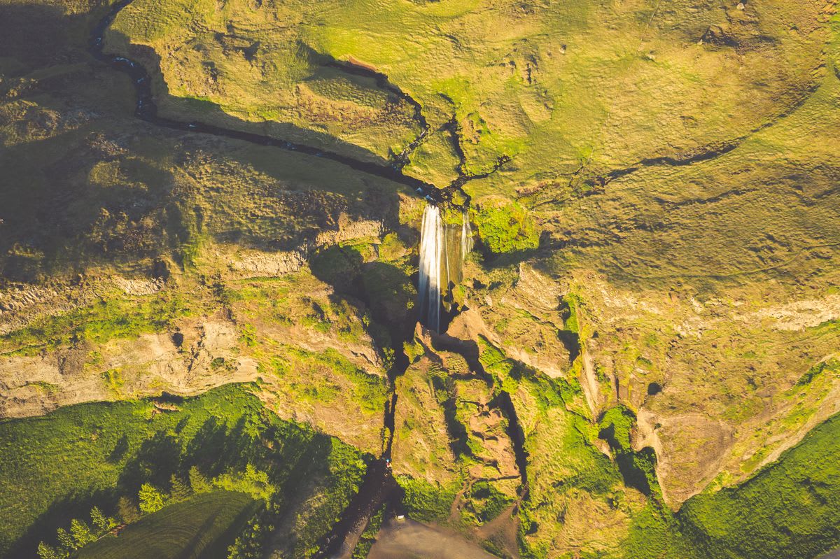 Aerial view of Seljalandsfoss waterfall cascading from green Iceland cliffs into a canyon, with winding river, trails, and golden light
