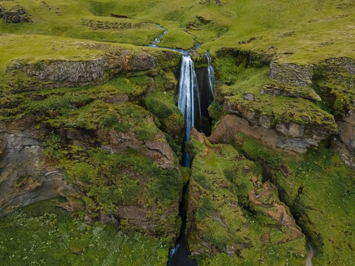 Drone view of Gljúfrabúi waterfall dropping into a narrow mossy Iceland canyon, surrounded by green cliffs, rocky trails, and streams