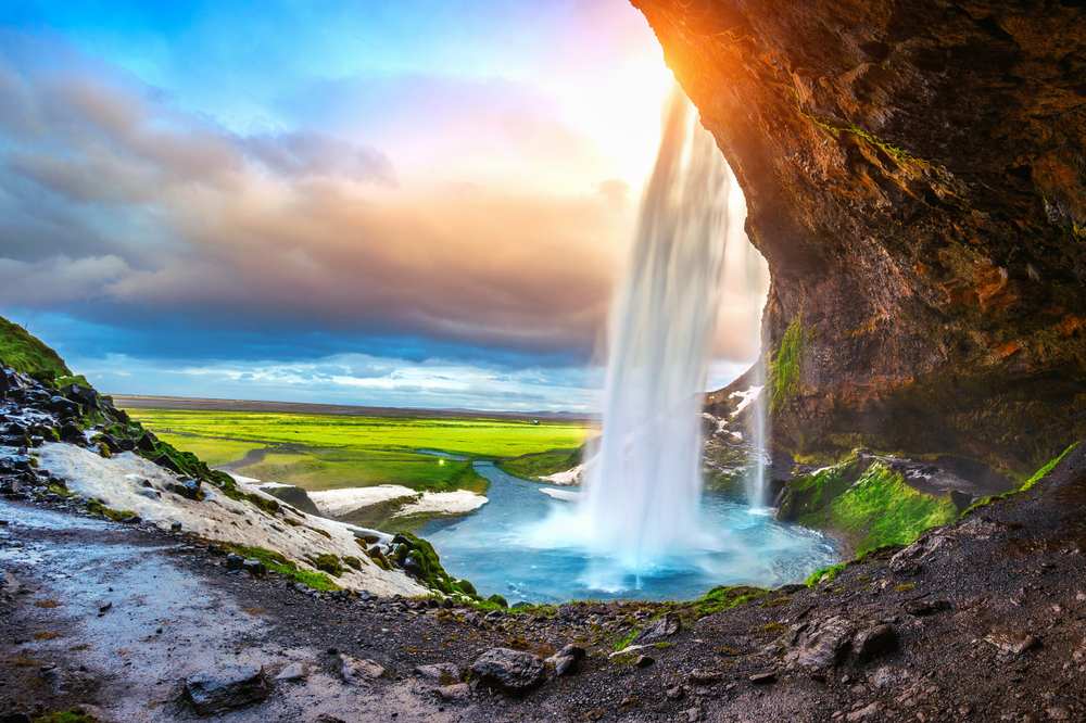 Behind Seljalandsfoss waterfall in South Iceland, sunset light streams through falling water over a rocky cave, blue pool, and green plains