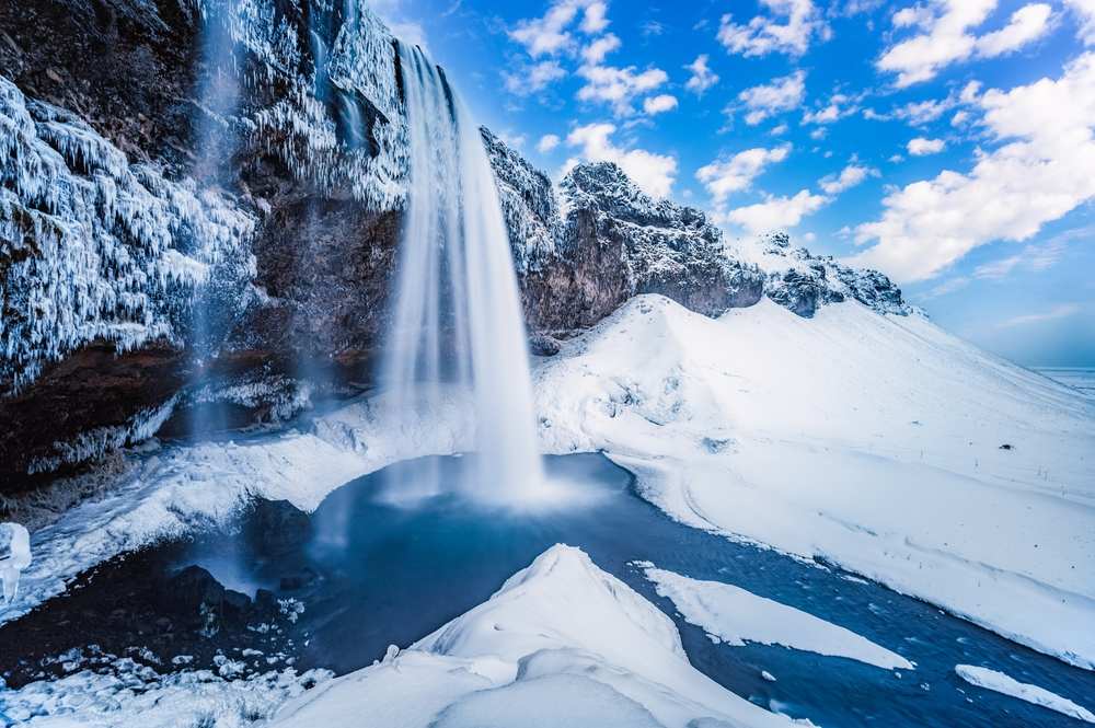 Winter view of Seljalandsfoss waterfall in South Iceland, cascading beside icy cliffs into a blue pool surrounded by snow and river