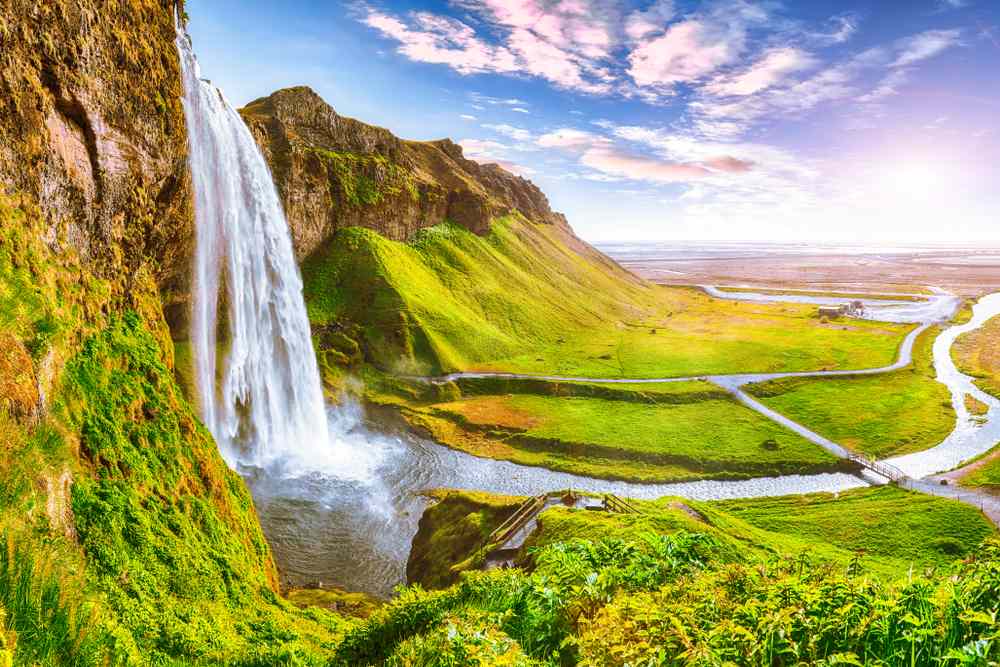 Summer view of Seljalandsfoss waterfall in South Iceland, pouring over mossy cliffs beside green hills, river trails, and bright sky