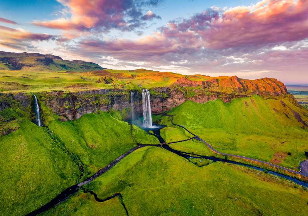 Aerial sunset view of Seljalandsfoss waterfall in South Iceland, with twin cascades, green cliffs, winding rivers, and dramatic pink clouds