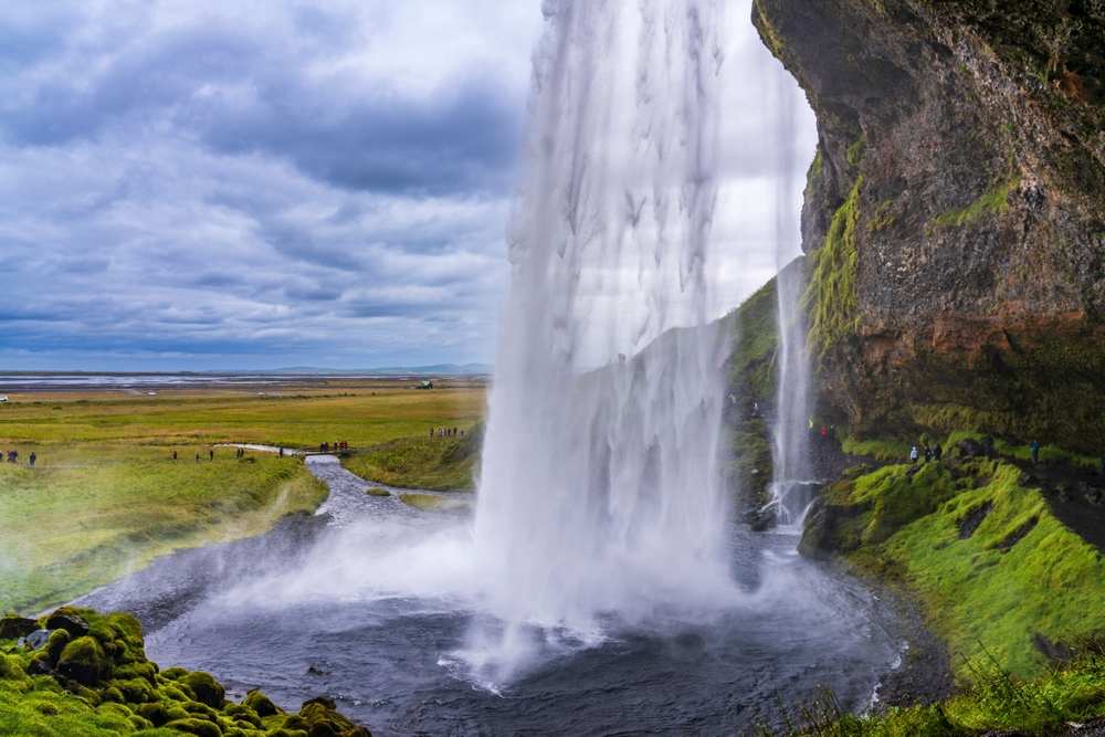 Close view behind Seljalandsfoss waterfall in South Iceland, with misty falls, mossy cliffs, blue pool, storm clouds, and green plains