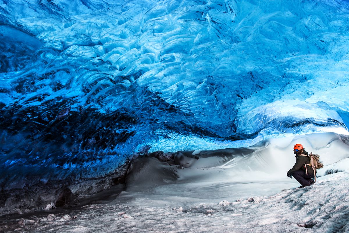 Hiker exploring a glowing blue ice cave beneath Vatnajökull glacier in Iceland, with frozen tunnel walls and snowy ground near Morsárfoss
