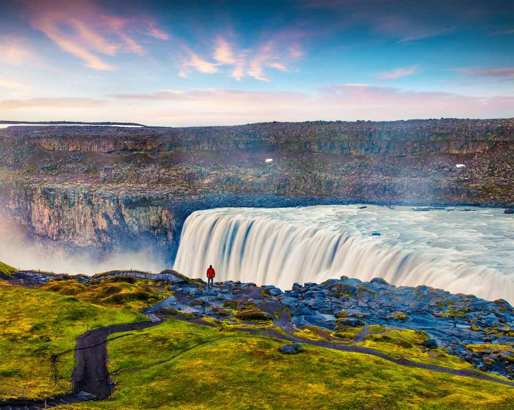Panoramic view of Dettifoss Waterfall in Iceland, with a red-jacketed traveler overlooking a misty basalt canyon under colorful sky
