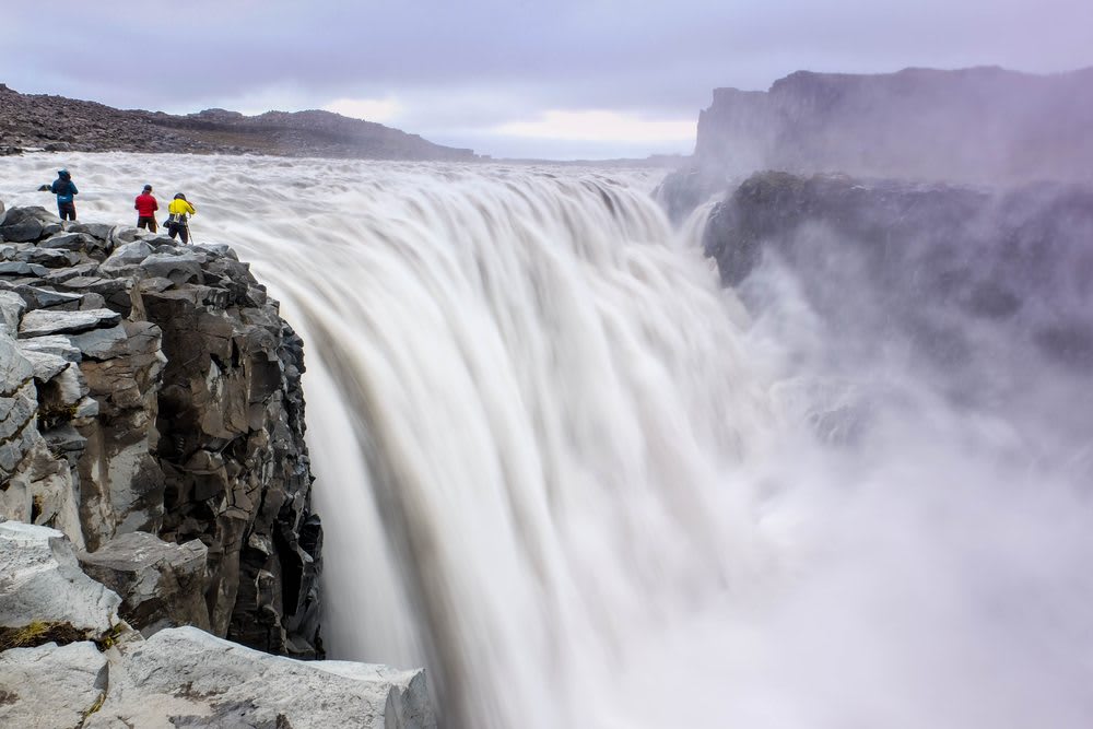 Hikers stand on rocky cliffs beside Dettifoss Waterfall in North Iceland as powerful glacial water plunges into a misty canyon