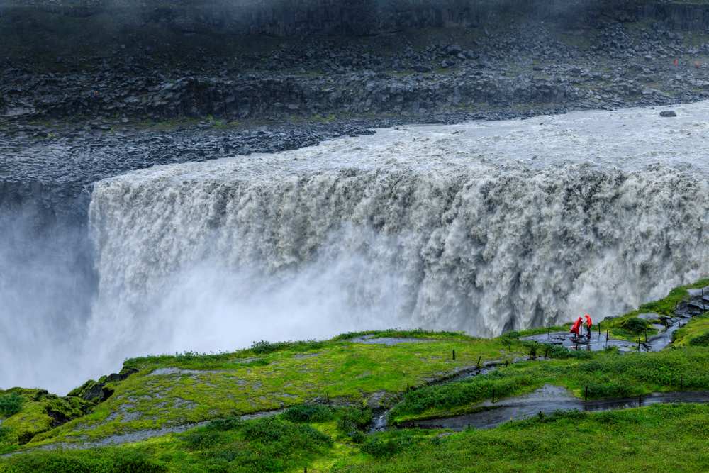 Dettifoss Waterfall thunders over basalt cliffs in North Iceland, with red-jacketed visitors on a green misty viewpoint trail